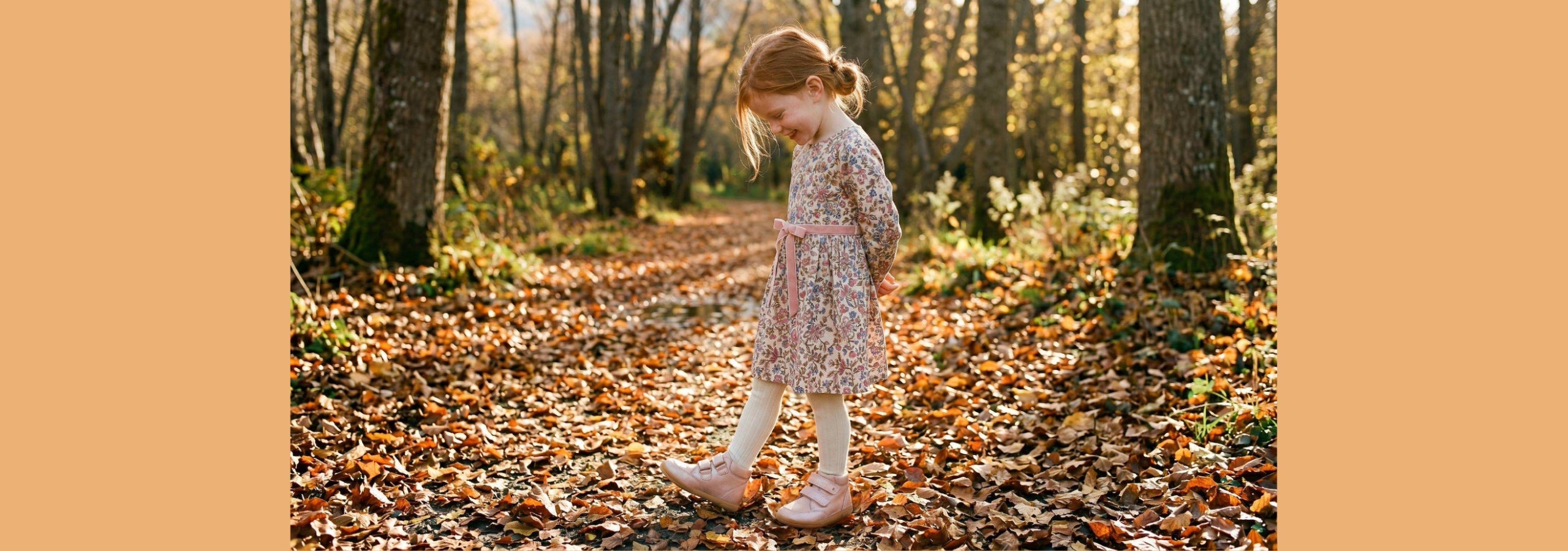 A young girl in a pink and white patterned dress walks along a leaf-covered path in a wooded area with autumn trees.