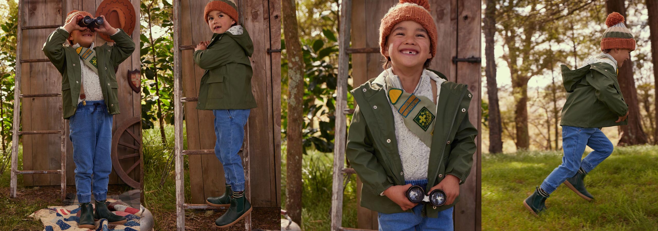 Child in a green jacket and blue jeans standing in front of a wooden structure with trees in the background.