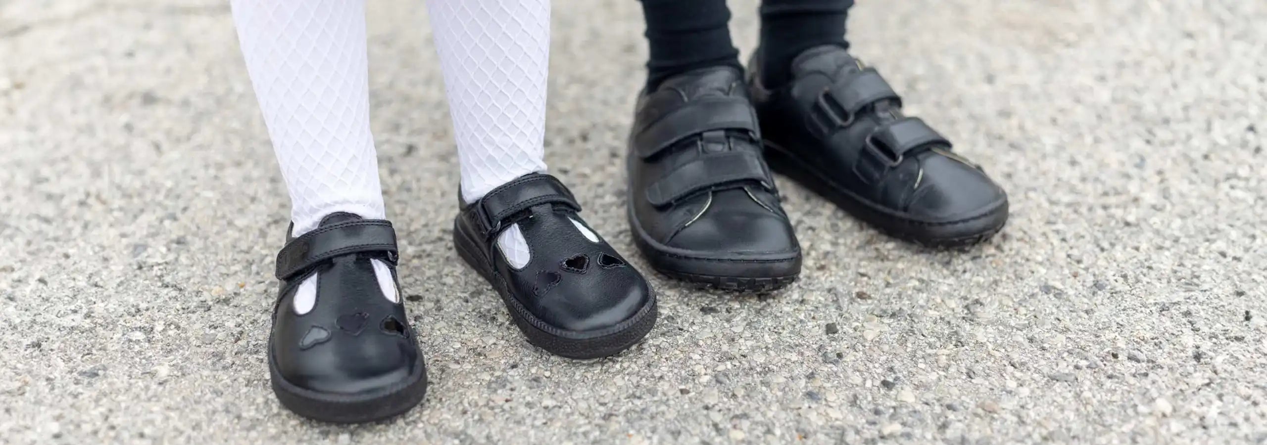 Three children's black shoes and black socks standing on a gray concrete ground.
